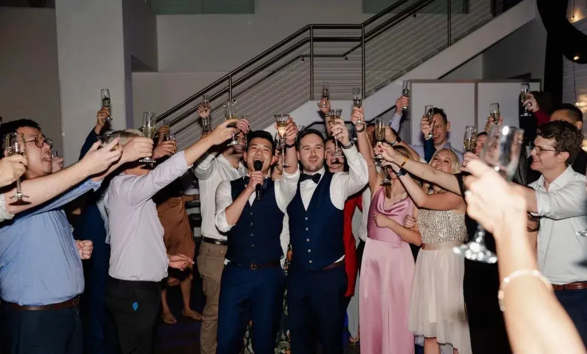 Two grooms leading a champagne toast surrounded by guests at a modern Southern California wedding venue
