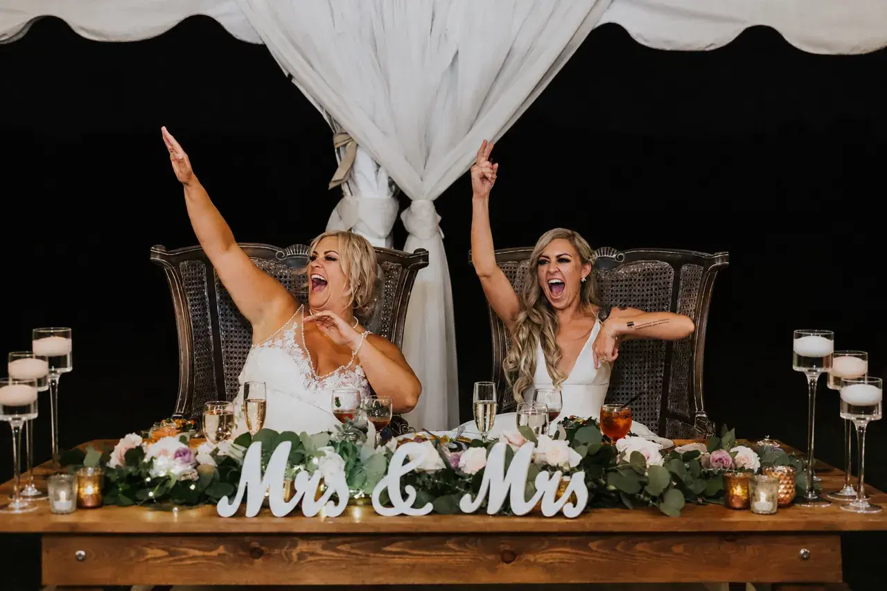 Two brides cheering with arms raised at their Mrs and Mrs sweetheart table during their wedding reception